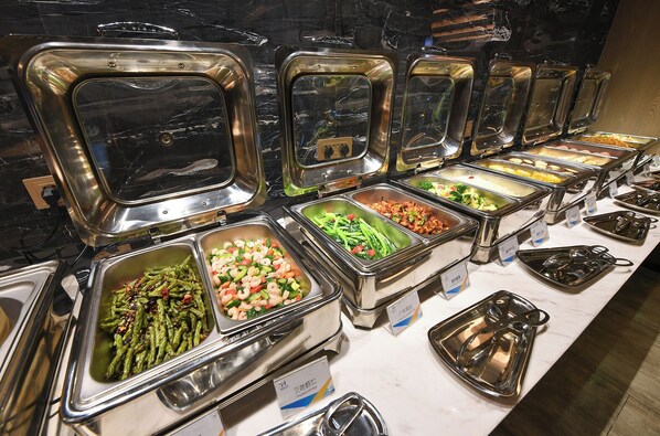 Un bufé con diversas opciones de comida en recipientes de acero inoxidable, dispuestas sobre una encimera de mármol.