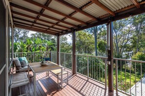 A covered outdoor deck with a wooden table and chairs, a potted plant, and a view of a lush garden.