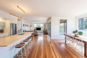A modern kitchen with a central island, bar stools, a dining area with chairs, and a large window.