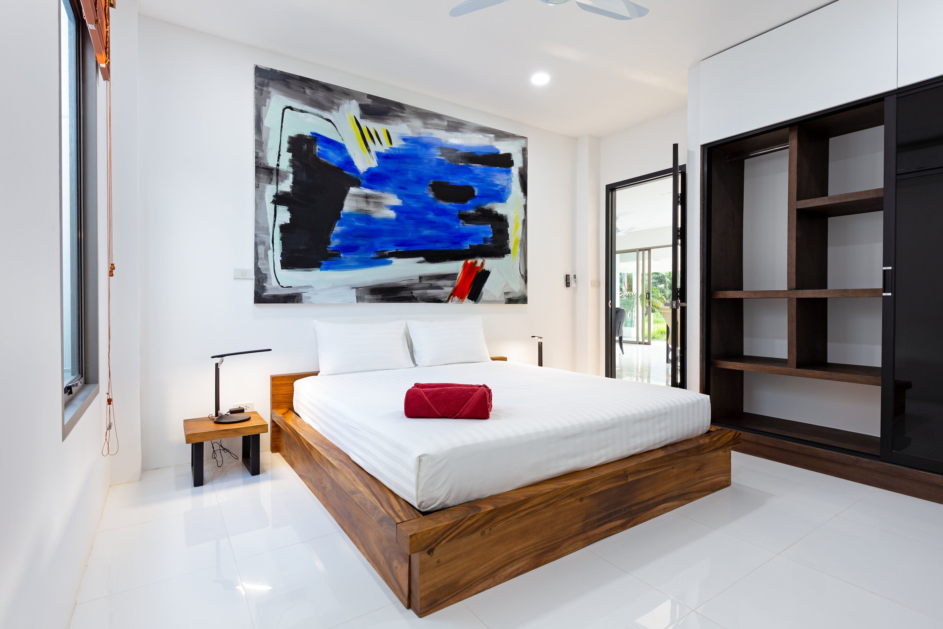 A modern bedroom with a large bed, a wooden headboard, a bedside table with a lamp, a large abstract painting on the wall, and a built-in shelving unit.