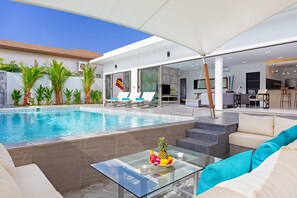 A modern outdoor pool area with lounge chairs, a glass table with fruit, and a patio with a sofa and coffee table. - Open-space Design 4BR Pool Villa Luna (Rawai)