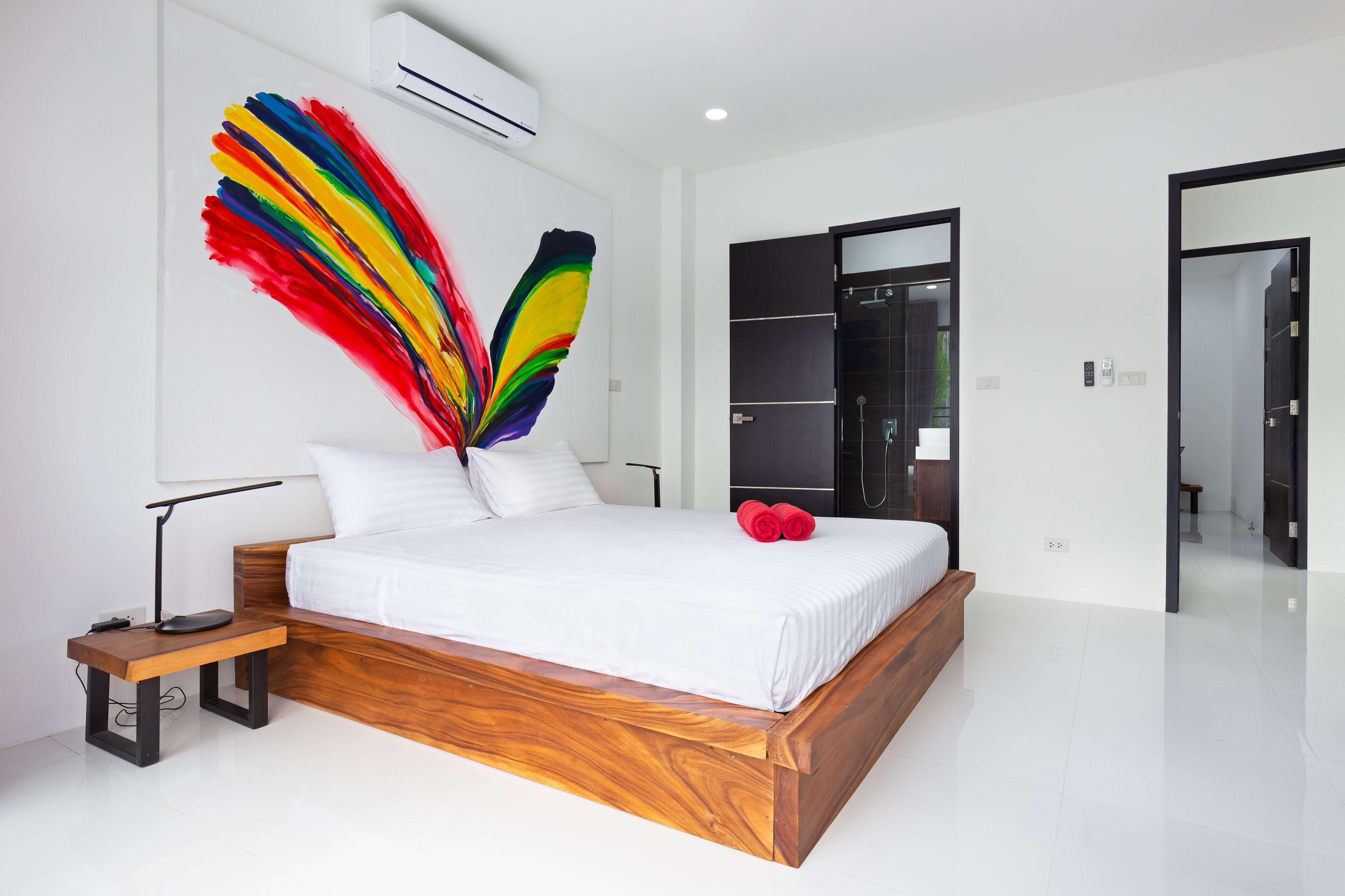 A modern bedroom with a wooden bed, a nightstand, a desk lamp, and a colorful feather artwork on the wall.