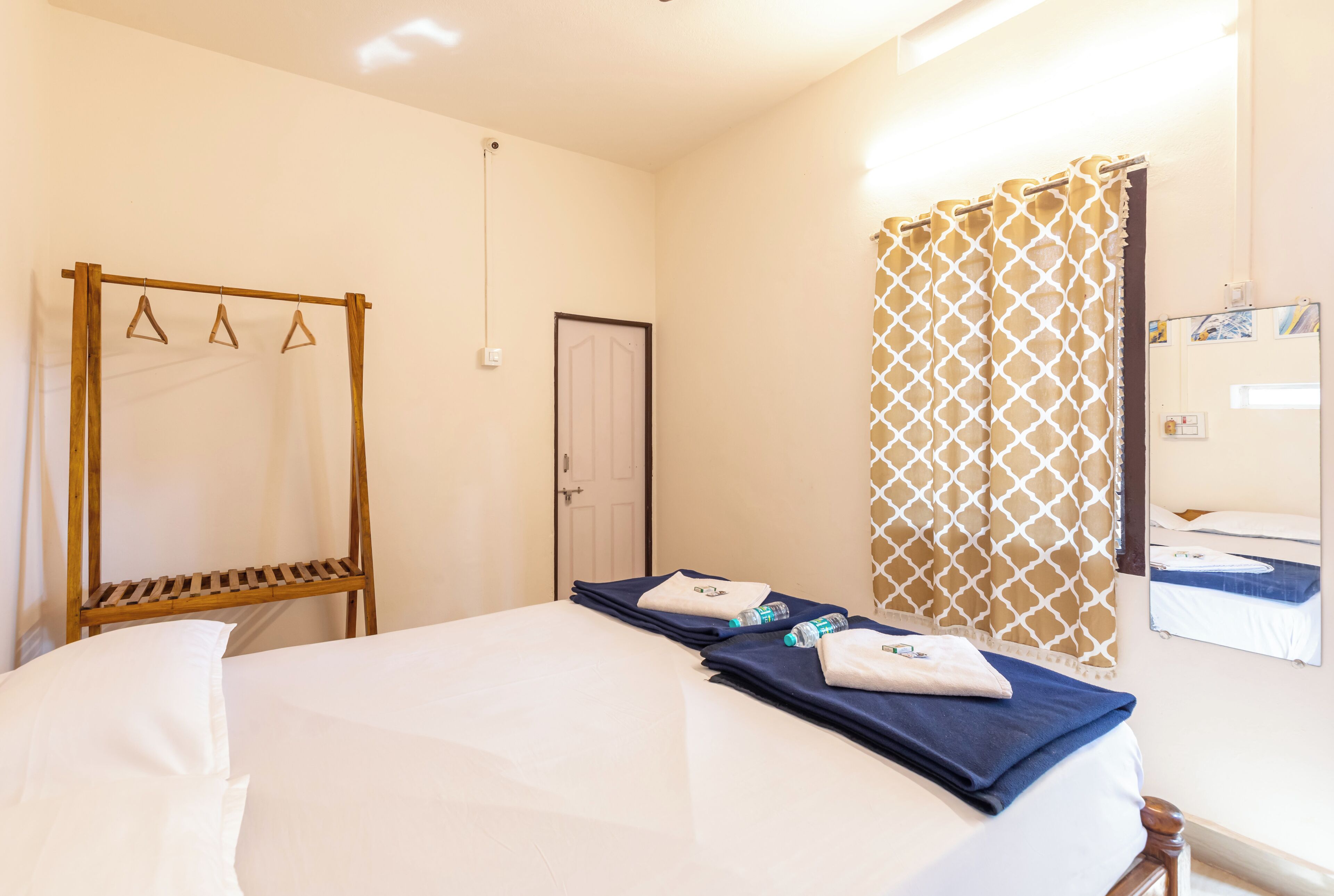 A hotel room with a bed, a wooden rack, a door, a patterned curtain, and a mirror reflecting a bed.