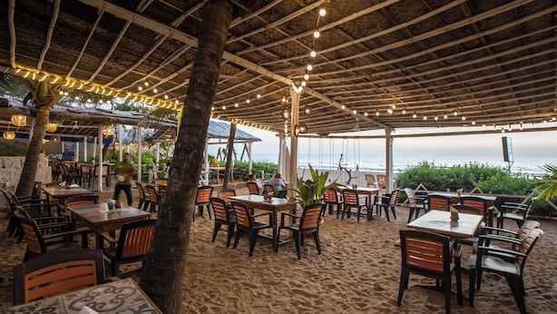 Un restaurante al aire libre con mesas, sillas, techo de paja e iluminacion con guirnaldas.