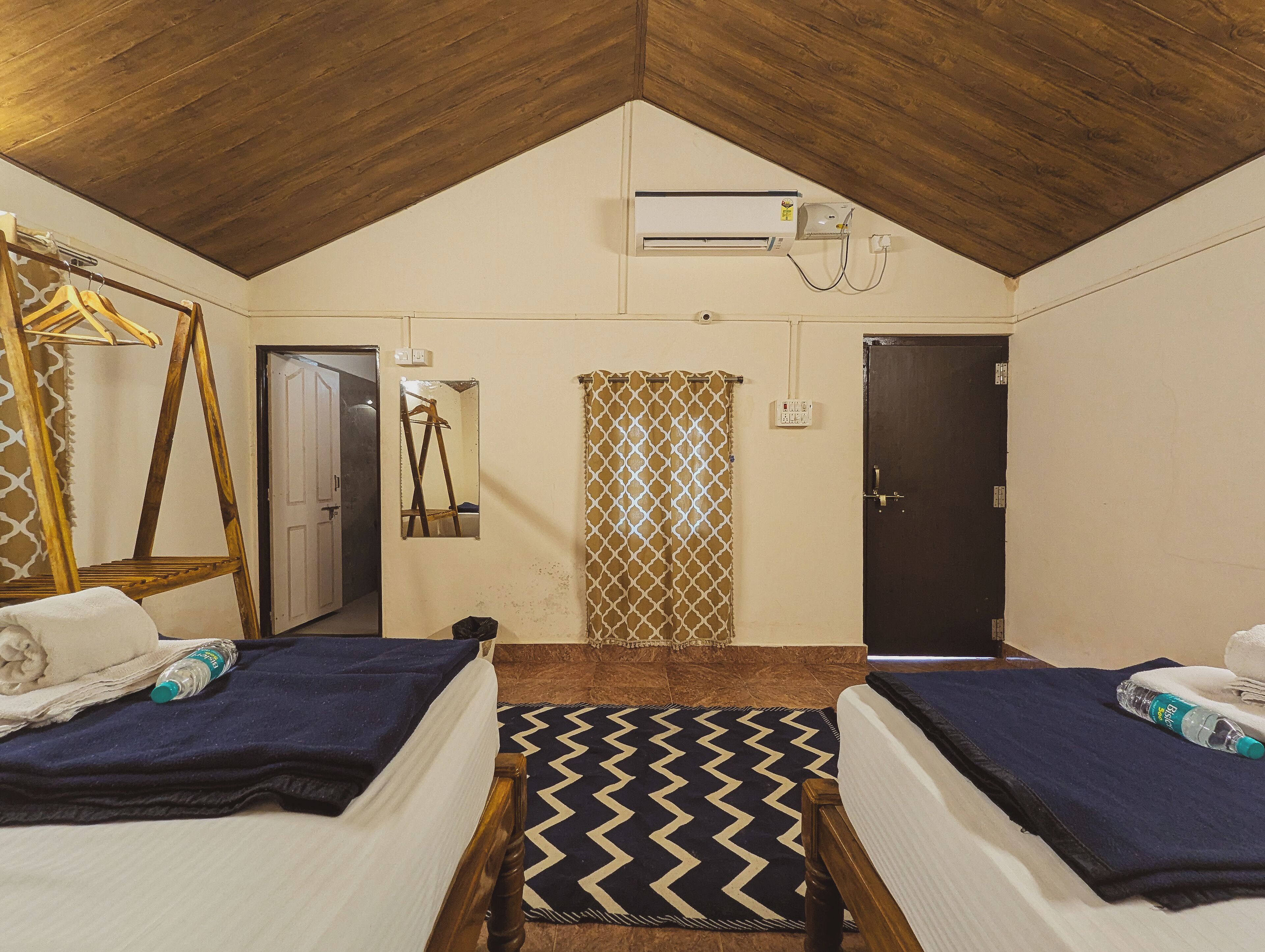 Een kamer met twee bedden, een houten plafond, een airconditioning, een spiegel, een deur en een kamerbreed gordijn.