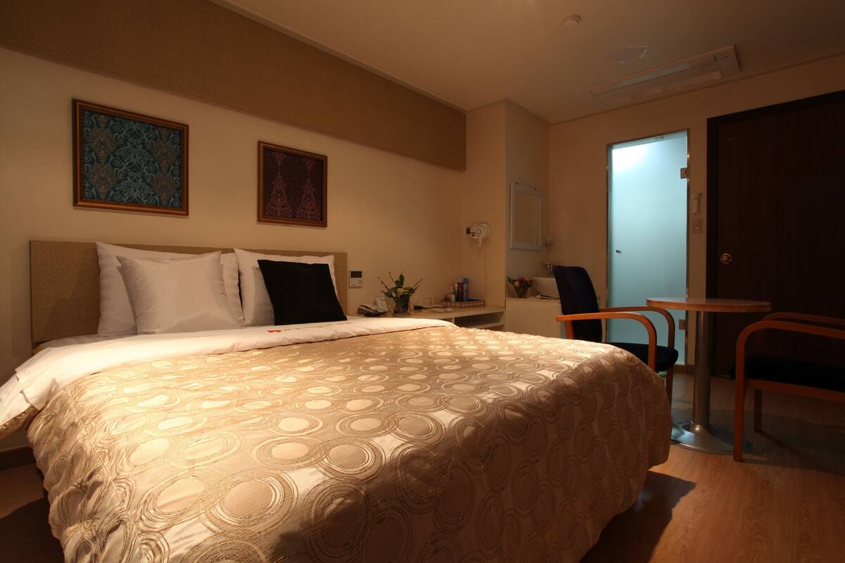 A hotel room with a large bed, a small desk, a chair, and a door.