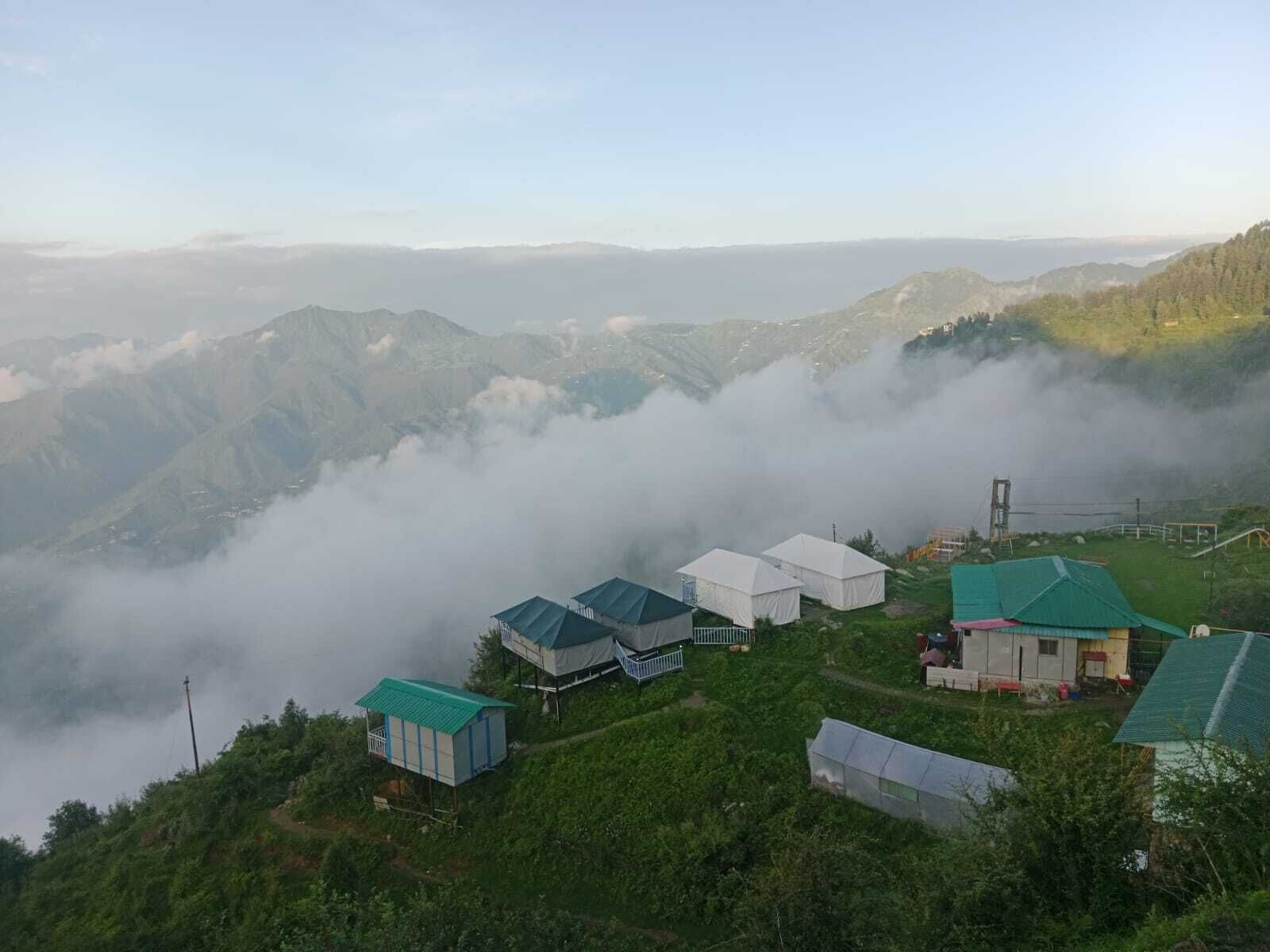 A mountainous landscape with several white tents and a few buildings with green roofs, surrounded by lush greenery and fog.