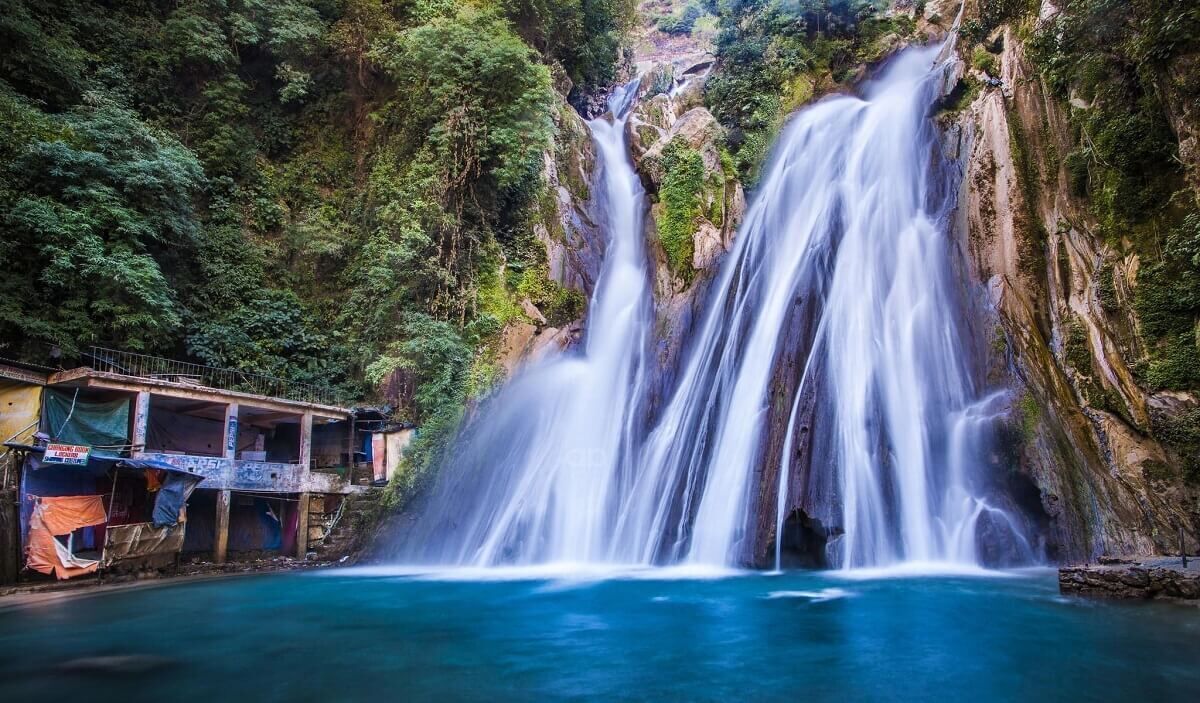 A waterfall cascading into a pool, surrounded by lush greenery and a building with a signboard.