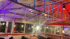 An indoor restaurant with tables and chairs, large windows, and hanging pendant lights. - Burans Breeze A Home Away From Home (Dhanaulti)