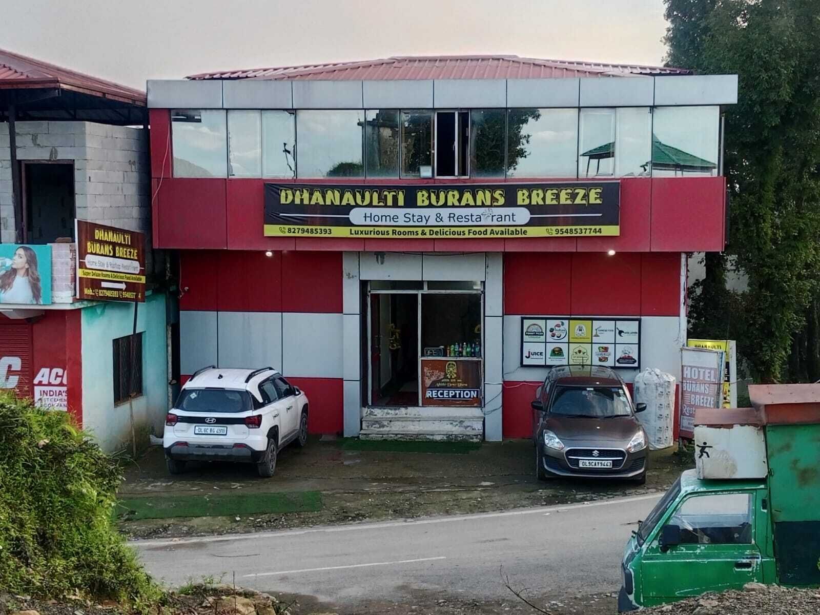 A building with a sign for 'Dhanavalli Burans Breeze' indicating a home stay and restaurant. There are two cars parked in front.