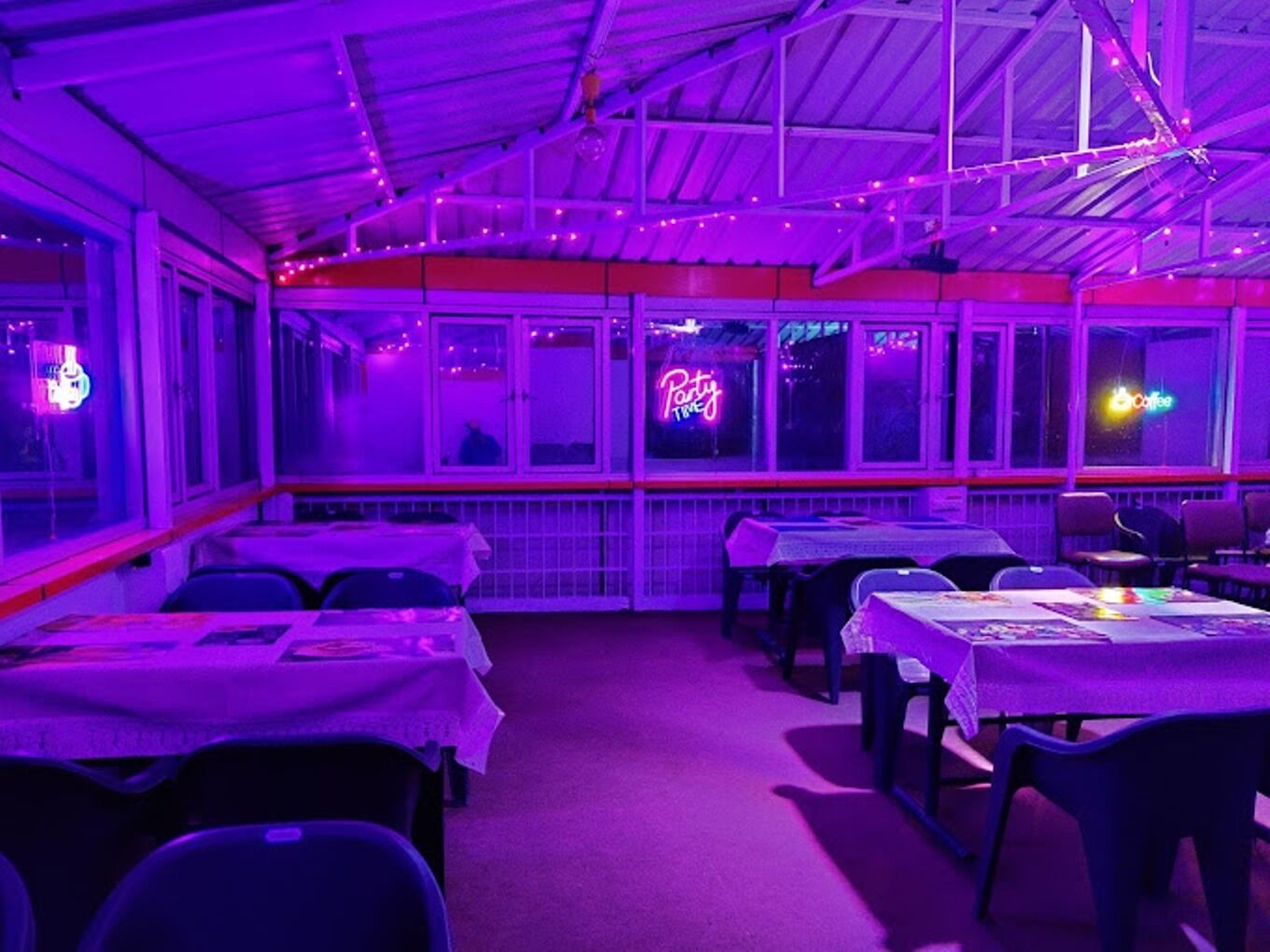 Un espacio interior para comer con mesas y sillas, un letrero de neón y una ventana con vistas.