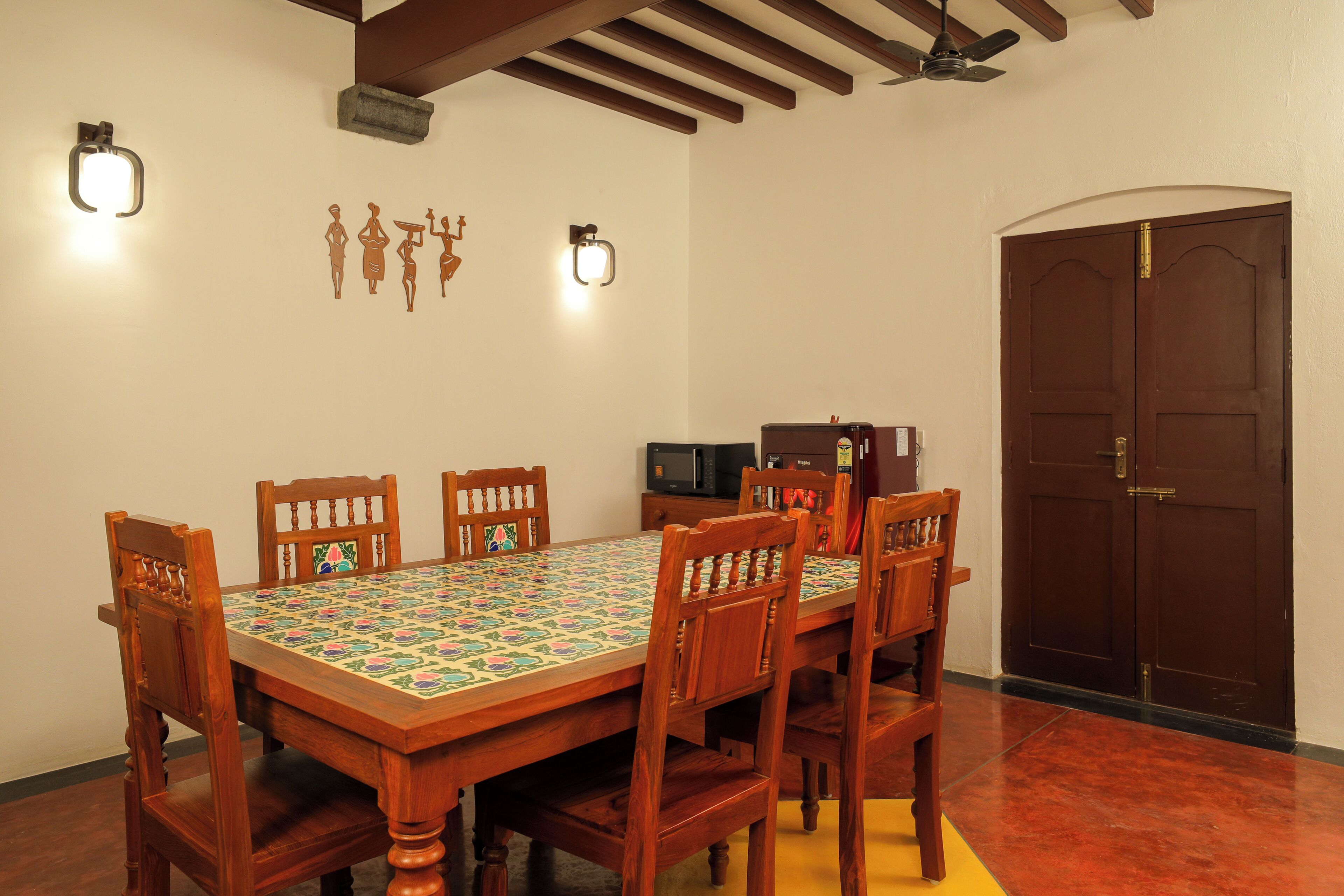 Un comedor con una mesa de madera, seis sillas, dos lámparas de pared, un microondas y una puerta cerrada.