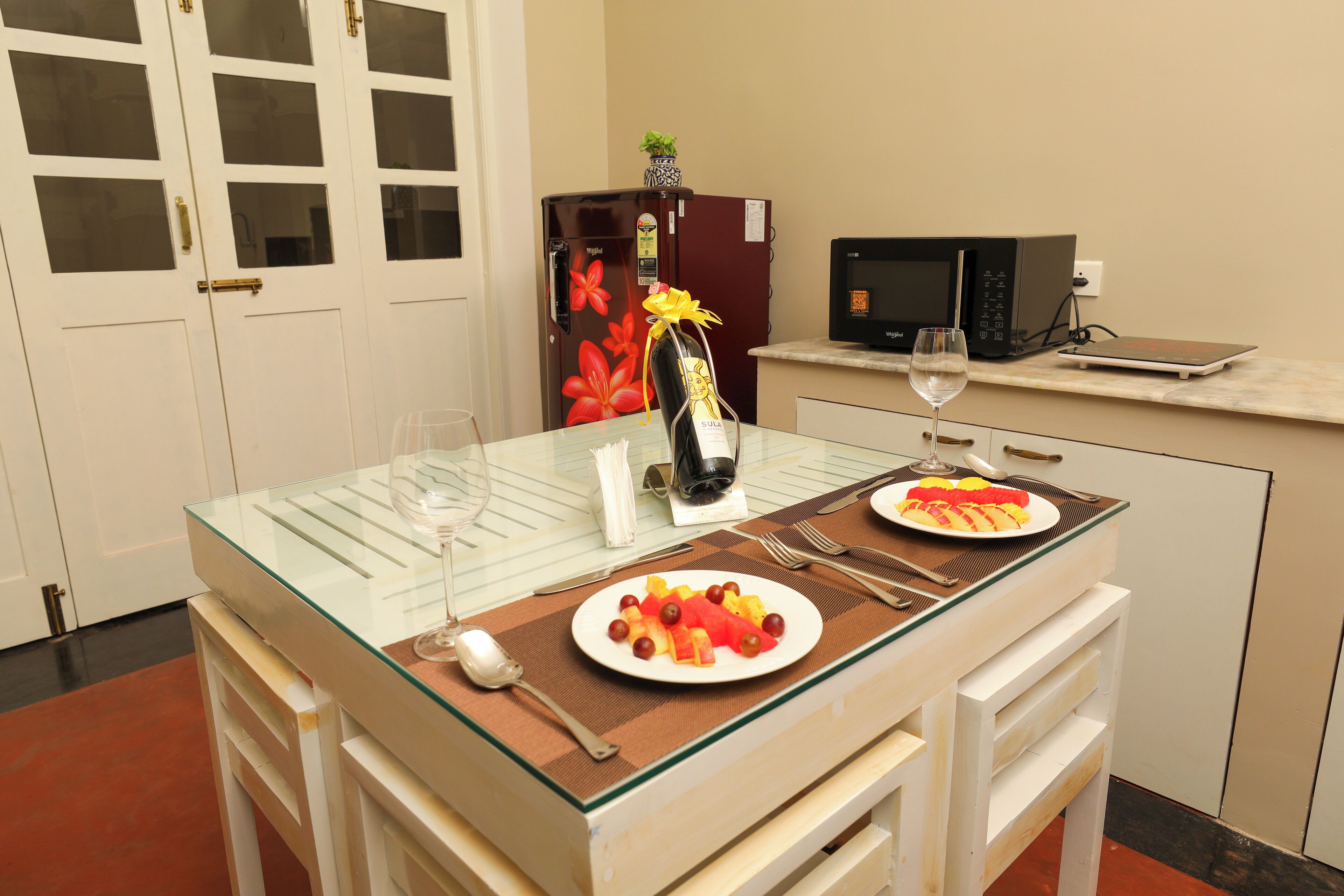 Un petit coin cuisine avec une table à plateau en verre, mise pour deux personnes, agrémentée d’une bouteille de vin, d’un verre et d’une assiette de fruits.