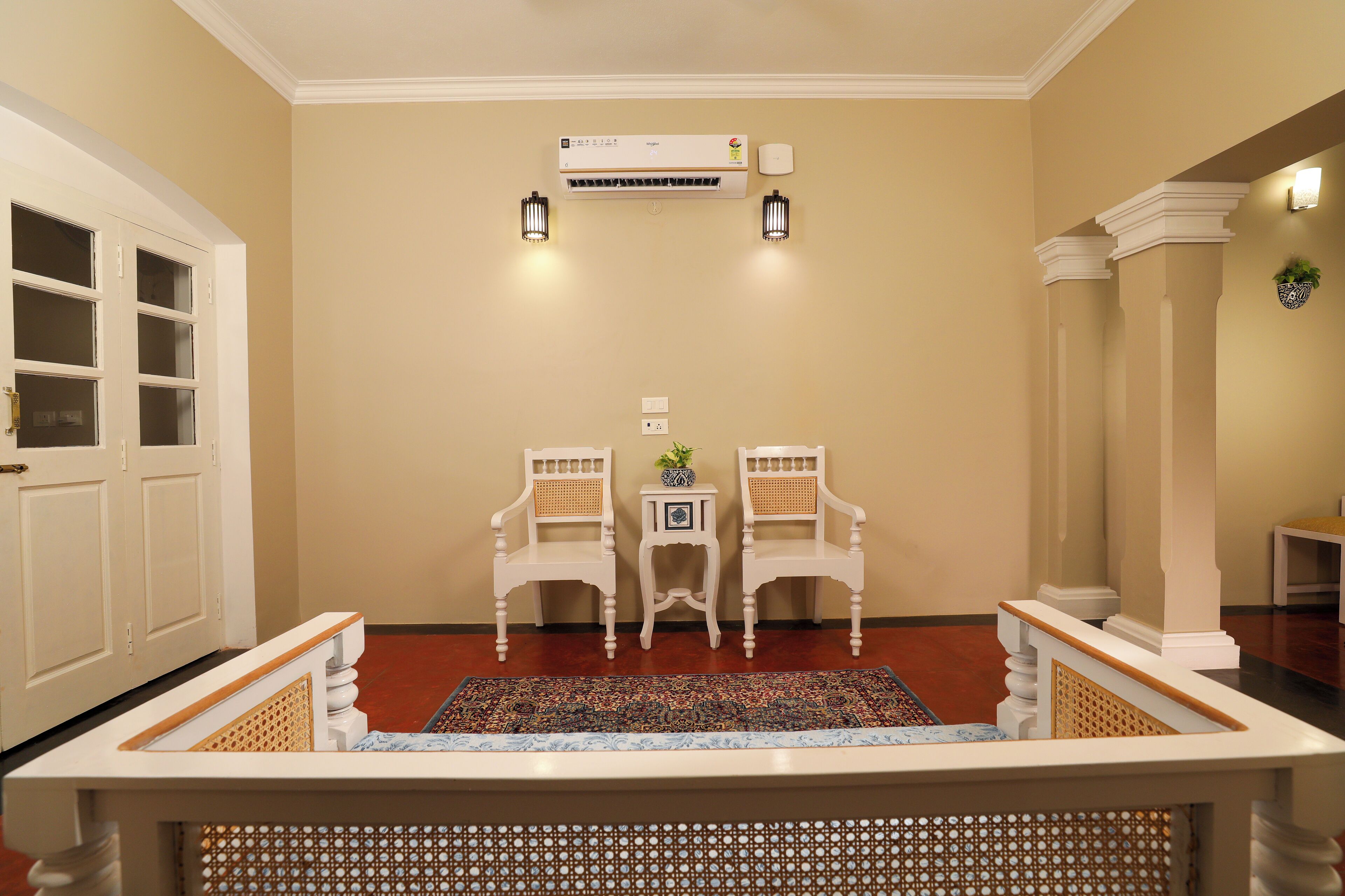A hallway with a beige wall, an air conditioning unit, two wall-mounted lights, a small table with a plant, and two white chairs.