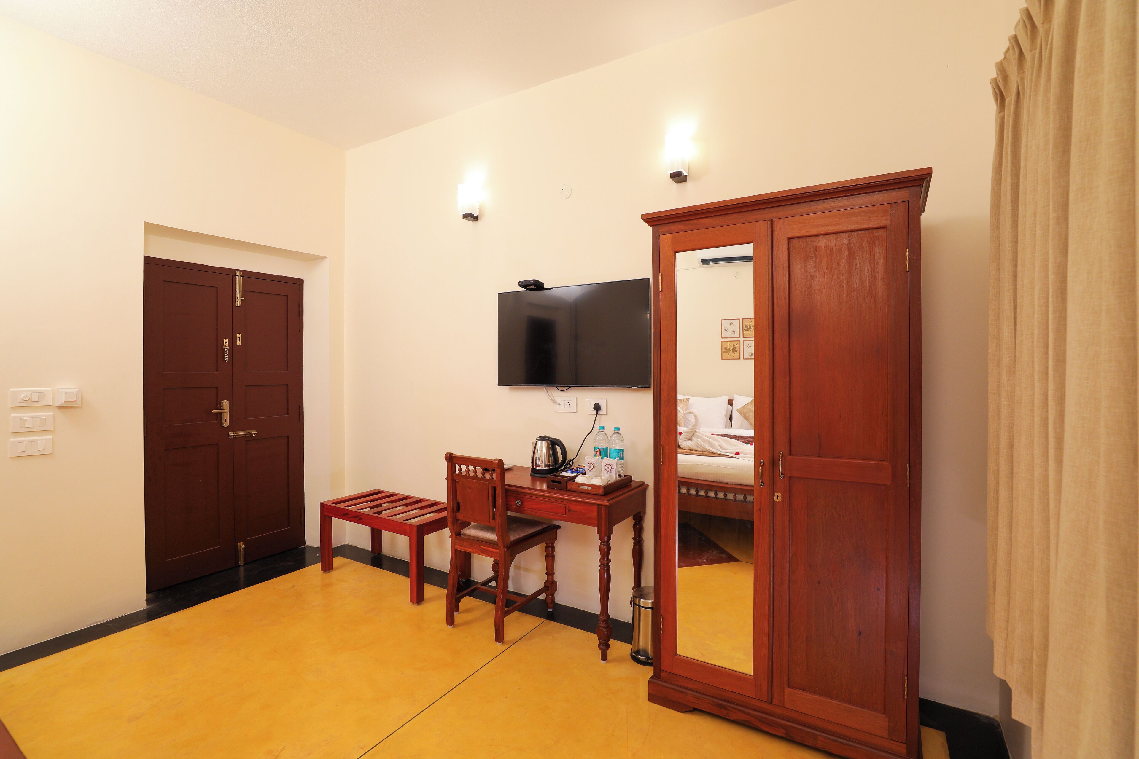 Une chambre d’hôtel avec un bureau, une chaise et une armoire en bois, un lit, un téléviseur fixé au mur et un miroir qui reflète une partie de la pièce.