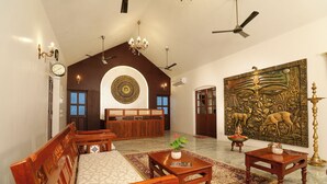 A spacious room with a high ceiling, wooden furniture, a large decorative wall piece, and a traditional rug. - FRANCO TAMIL VILLAS (Puducherry)