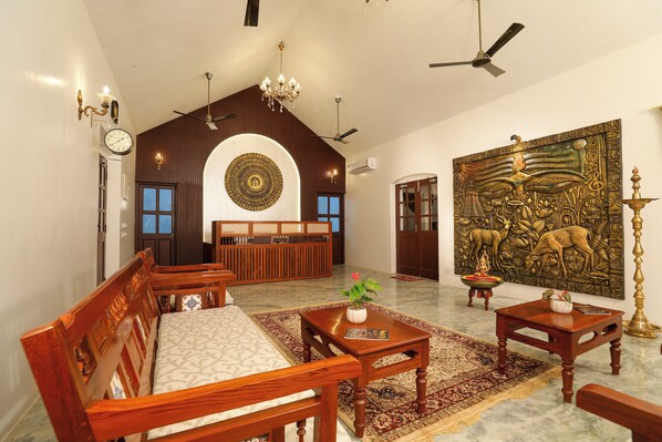 A spacious room with a high ceiling, wooden furniture, a large decorative wall piece, and a traditional rug. - FRANCO TAMIL VILLAS (Puducherry)
