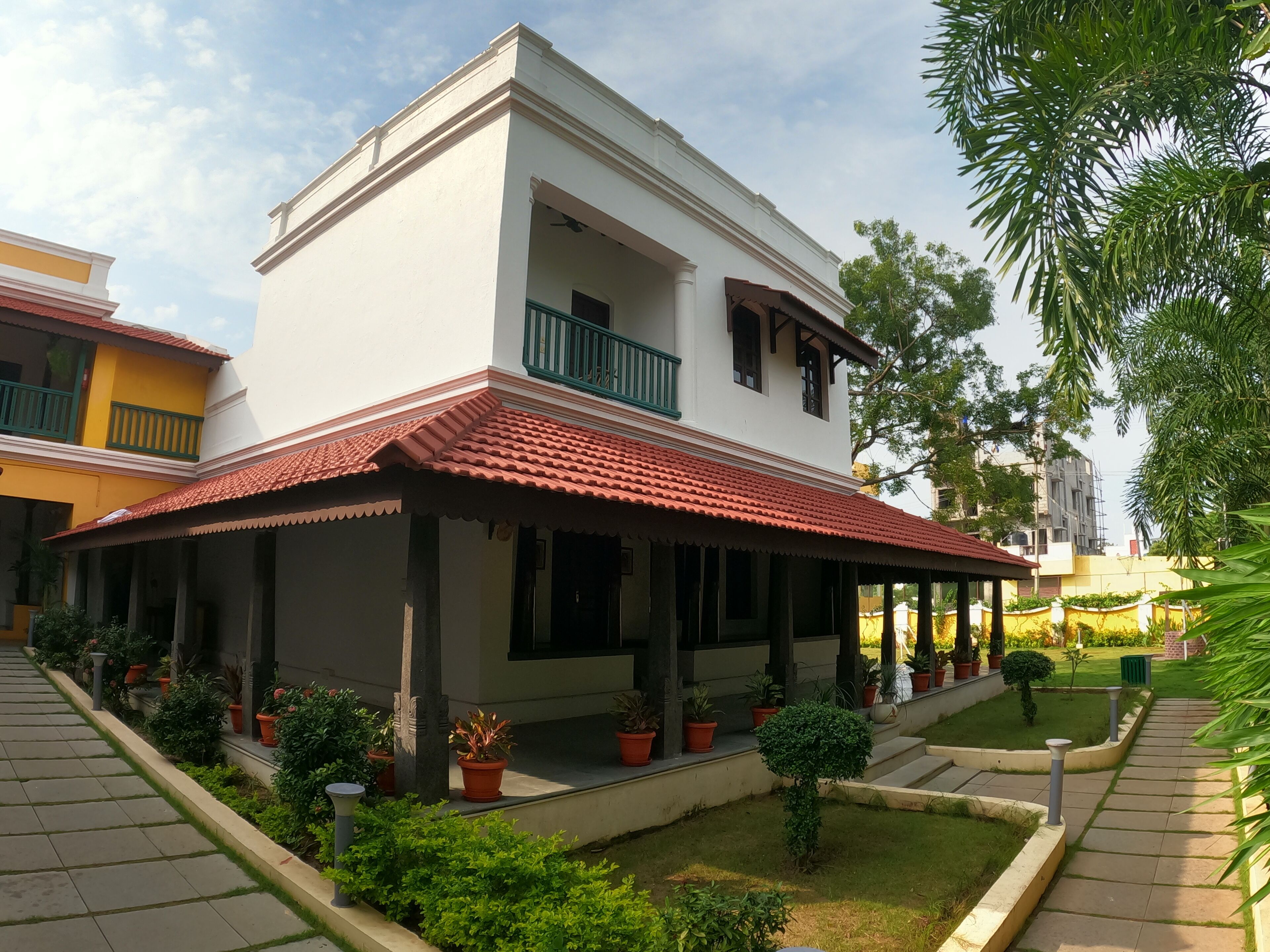 A two-story building with a red-tiled roof, a balcony with green railings, and a garden area with potted plants.