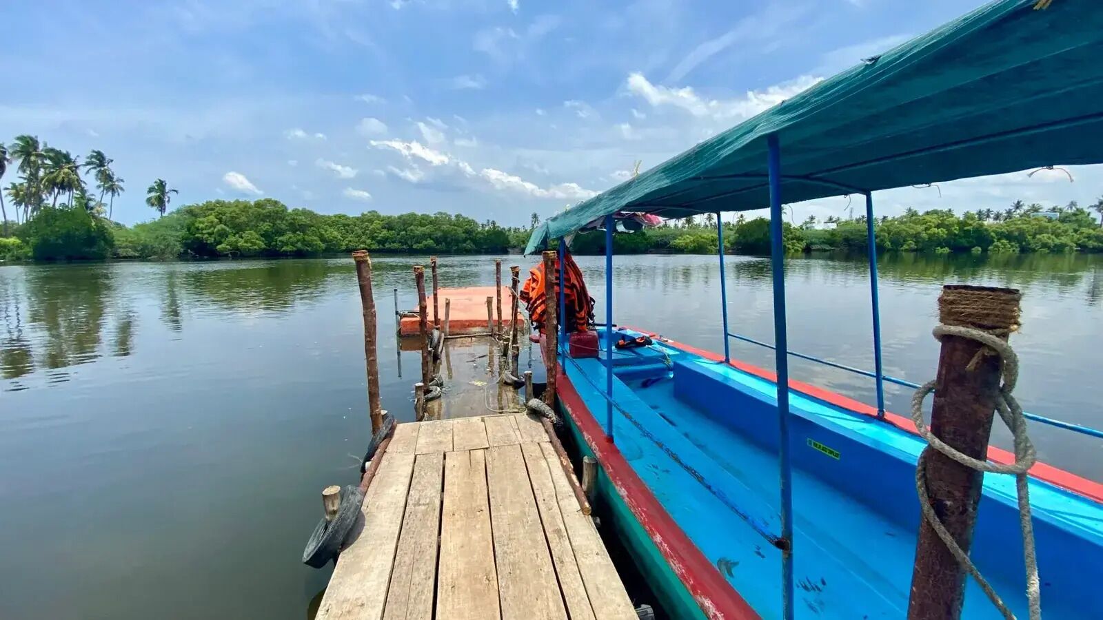 A boat docked at a wooden pier with a blue and red railing, surrounded by calm water and greenery.