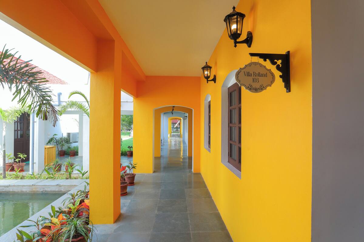 A corridor with yellow walls, a sign reading "Villa Roland 103", and potted plants along the sides.