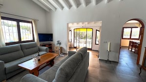 A modern living room with a grey sofa, a wooden coffee table, and a TV. There is a dining area with a wooden table and chairs, and a hallway leading to another room. - Beautiful and comfortable villa in Calpe, Costa Blanca, Spain  with private pool for 4 persons (Calpe)