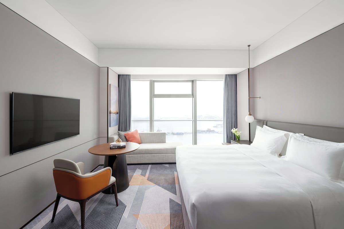 Une chambre d’hôtel moderne dotée d’un grand lit, d’une télévision à écran plat, d’un coin salon avec un canapé et des fauteuils, et offrant une vue sur la ville grâce à de grandes fenêtres.