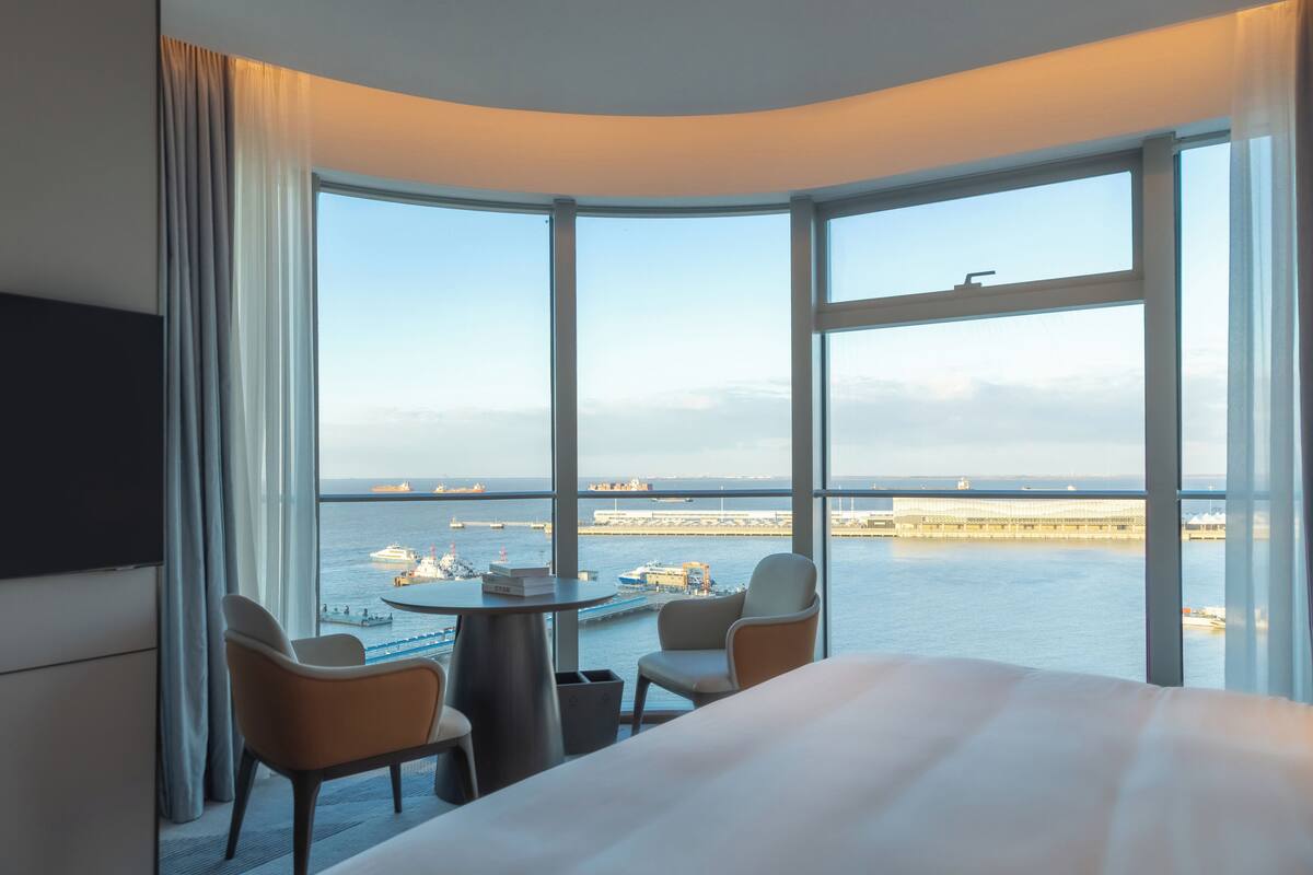 Une chambre d’hôtel avec une fenêtre incurvée, un lit, deux chaises, une petite table et un téléviseur.