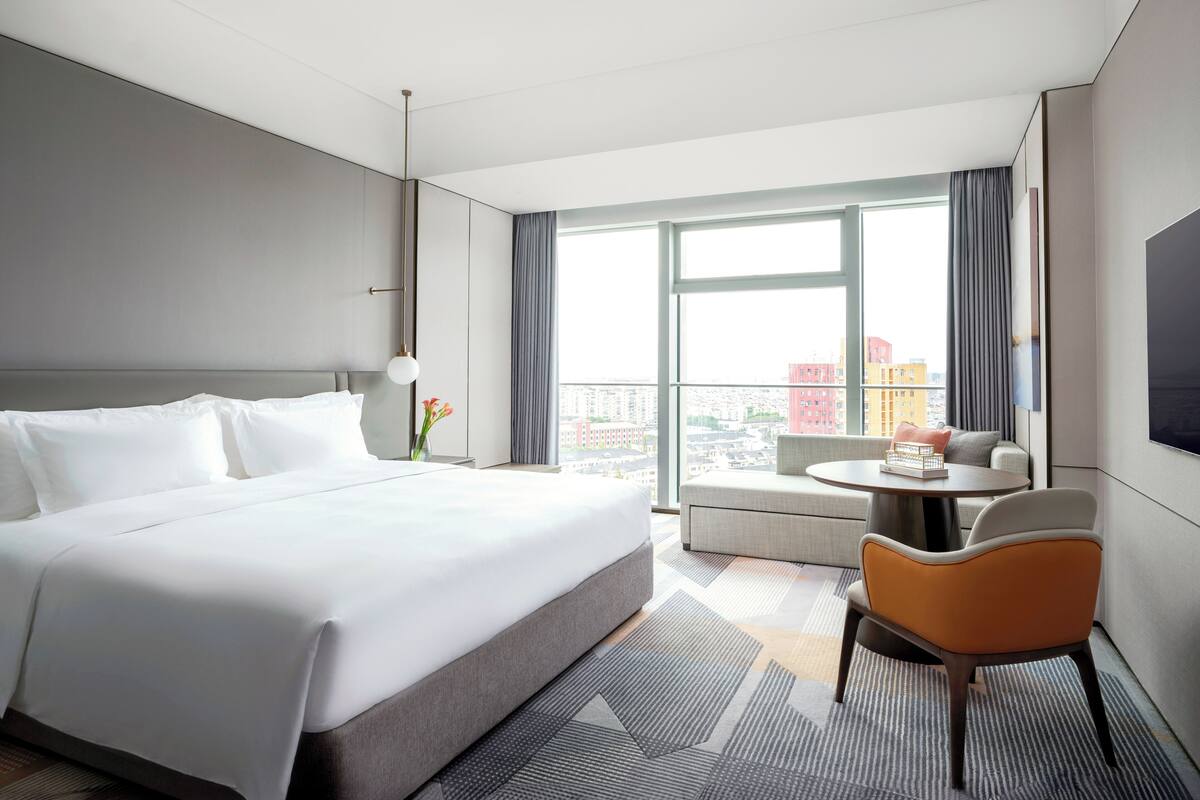Une chambre d’hôtel moderne dotée d’un grand lit, d’un coin salon avec une table et offrant une vue sur la ville grâce à de grandes fenêtres.