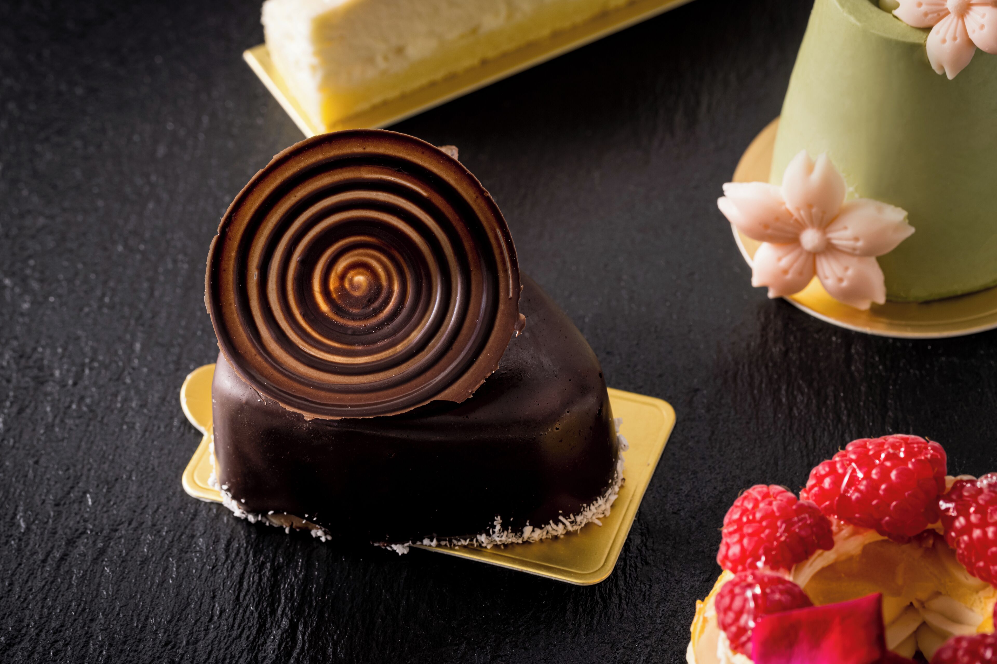 A chocolate-covered dessert with a spiral design on a golden plate, accompanied by a green cake with floral decorations and a fruit-topped pastry.
