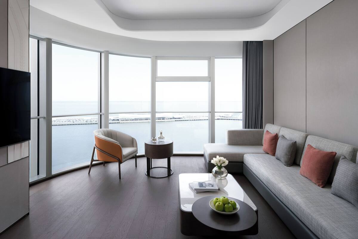 Une chambre d’hôtel moderne dotée d’une grande fenêtre donnant sur la mer, d’un canapé, d’un fauteuil, d’une table basse et d’un téléviseur à écran plat.