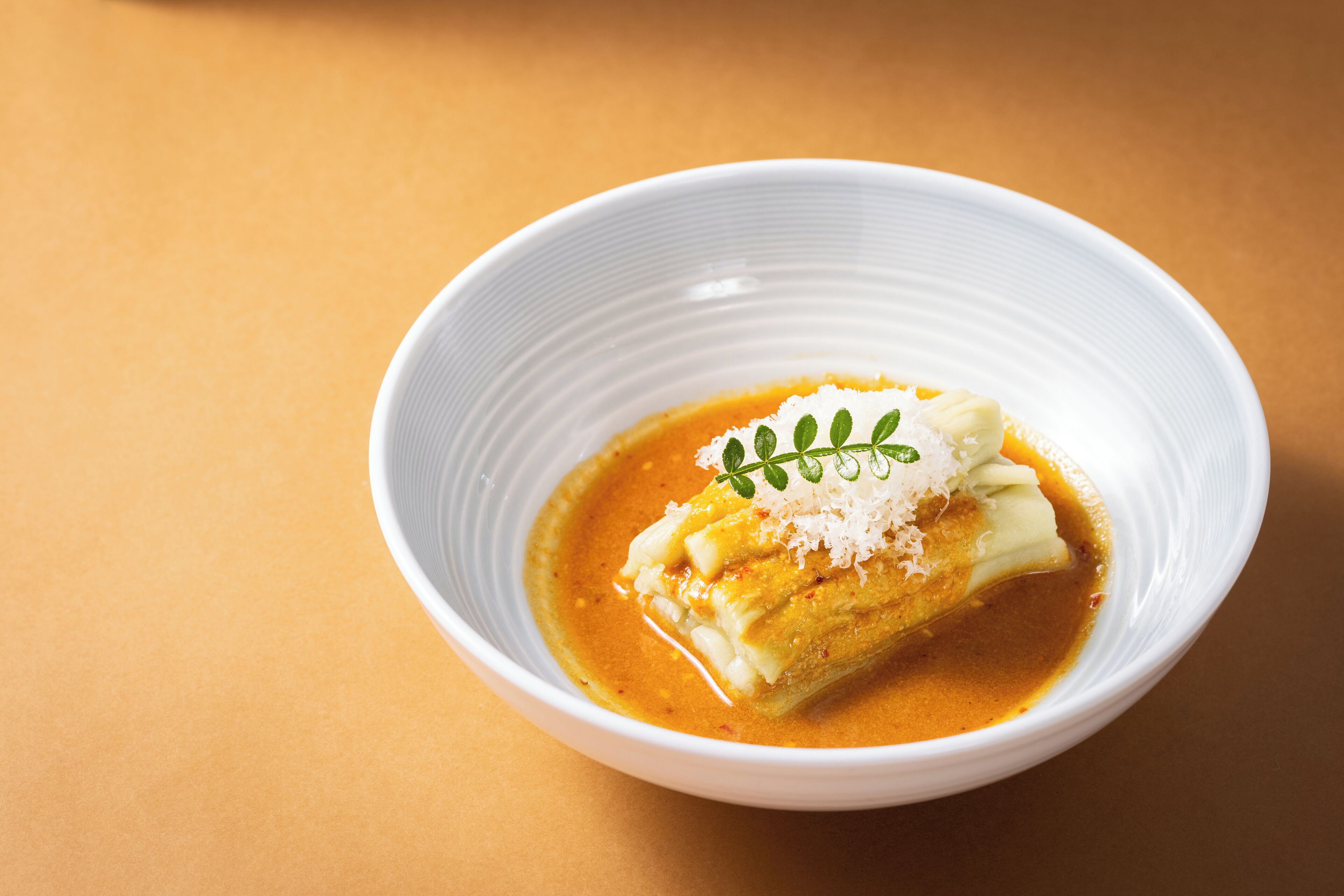 Un bol blanc contenant une assiette sur laquelle se trouve un aliment enroulé, peut-être une pâtisserie, recouvert de fromage râpé et agrémenté d’une garniture verte, le tout sur un fond orange uni.