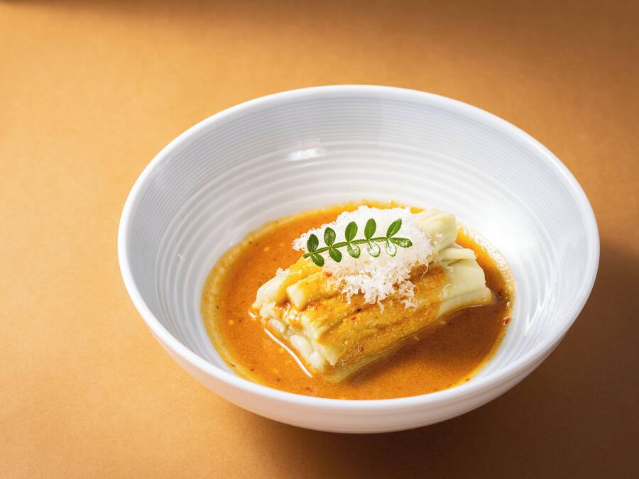 Un bol blanc contenant une assiette sur laquelle se trouve un aliment enroulé, peut-être une pâtisserie, recouvert de fromage râpé et agrémenté d’une garniture verte, le tout sur un fond orange uni.
