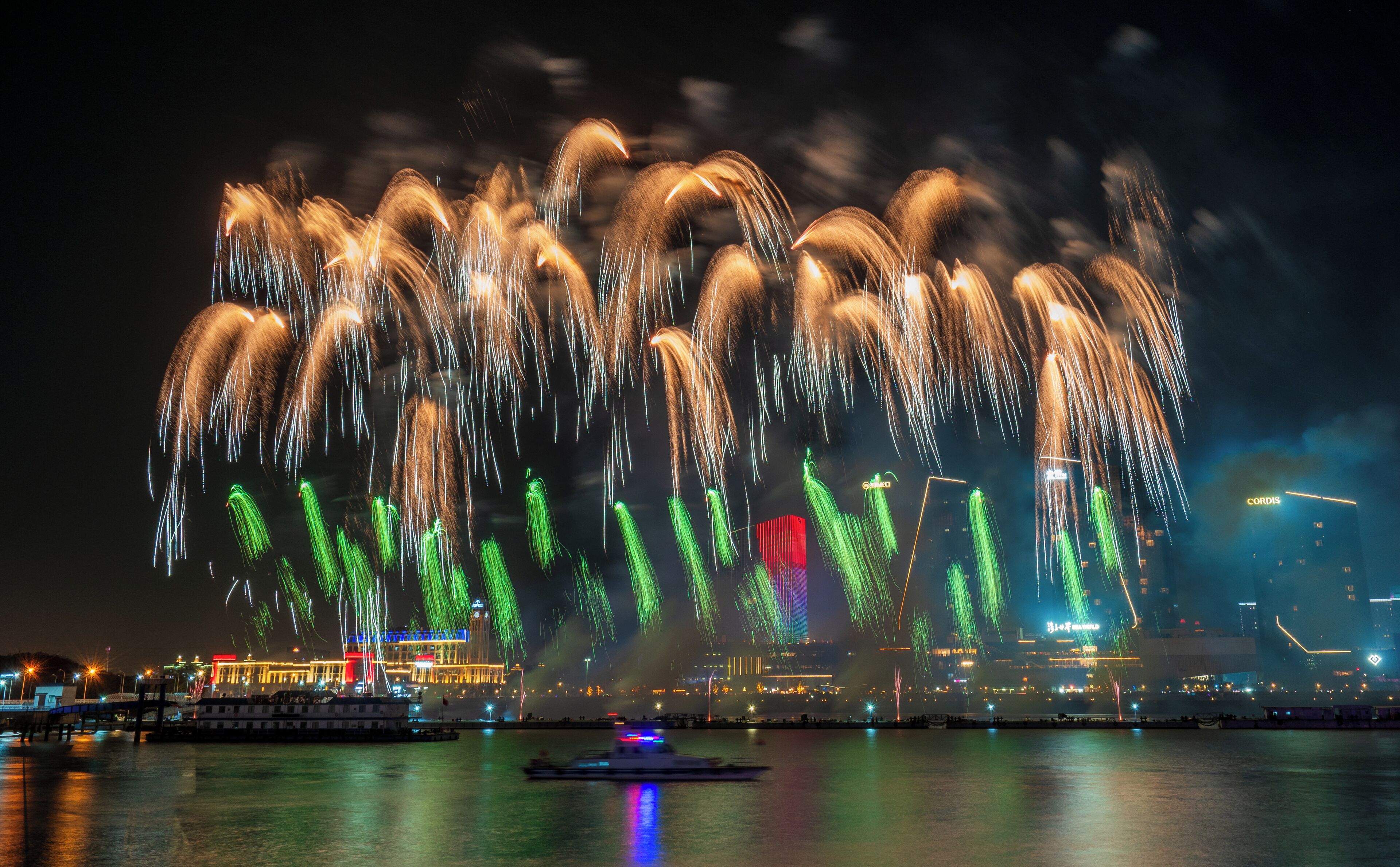 A waterfront view with fireworks display, boats, and illuminated buildings.