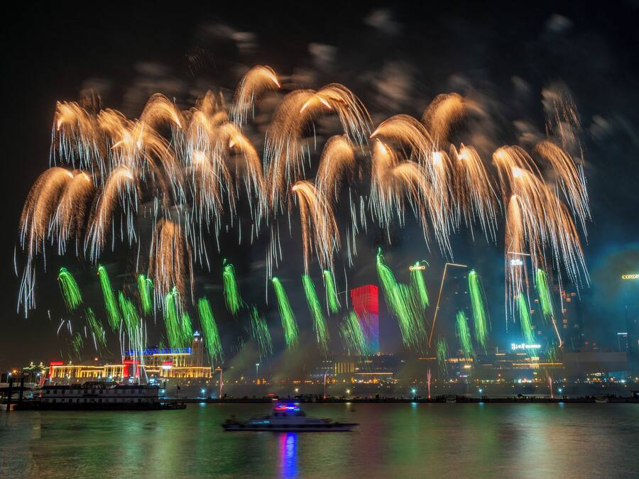 A waterfront view with fireworks display, boats, and illuminated buildings.