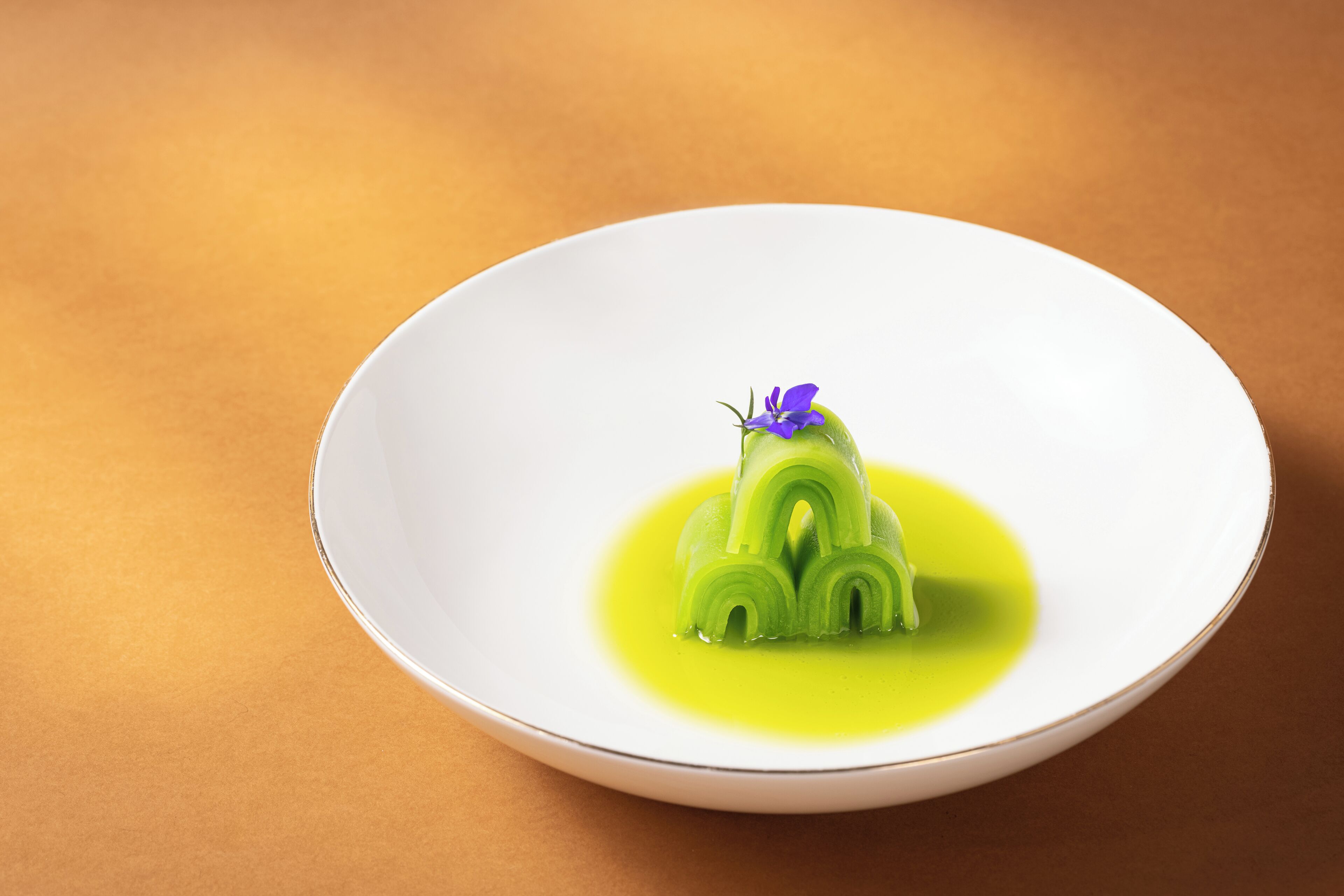 Une assiette blanche sur laquelle on voit un dessert vert et une fleur violette, le tout posé sur une surface marron.