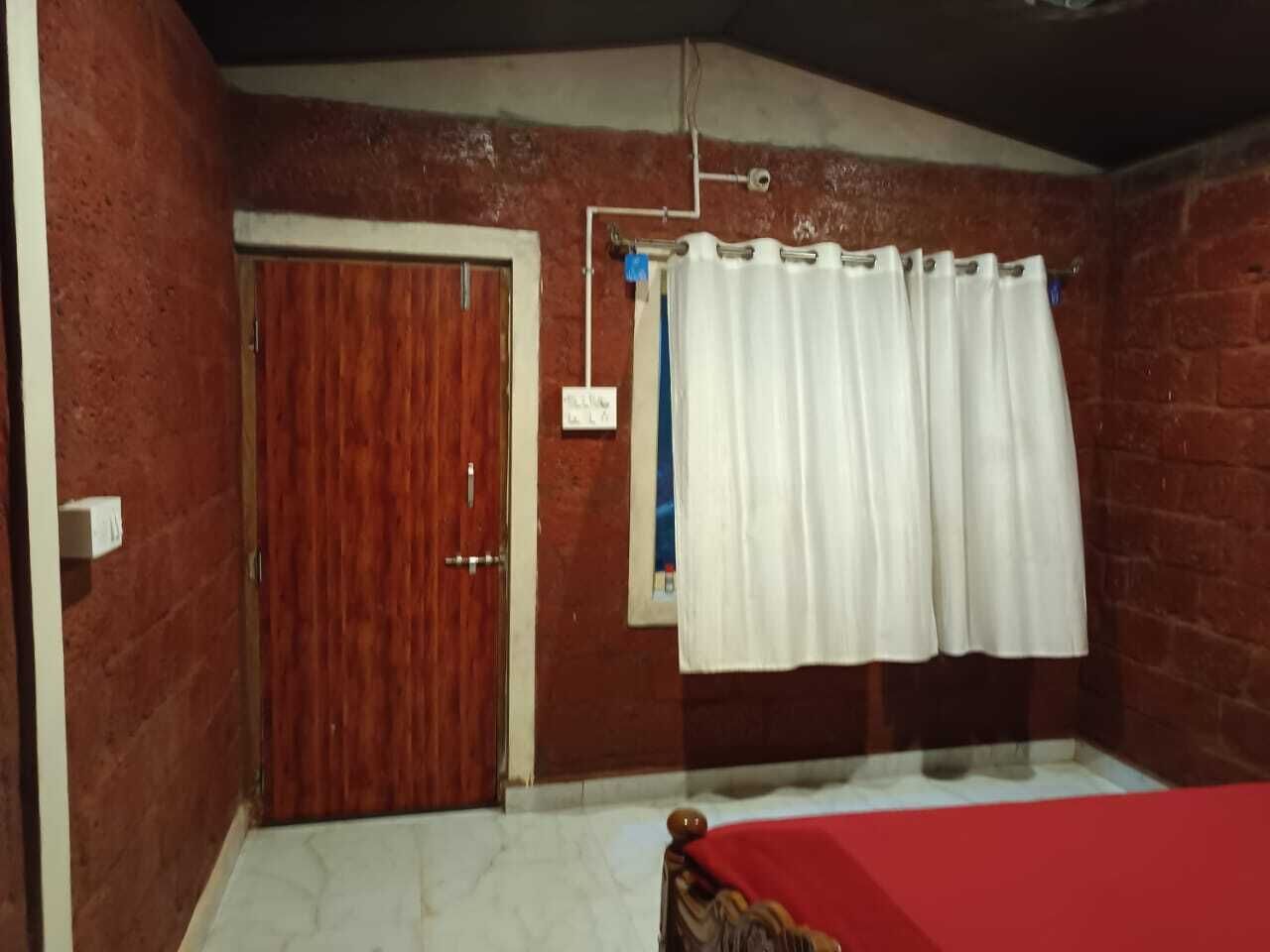 Ein Zimmer mit Holztür, weißen Vorhängen und einer roten Bettdecke.