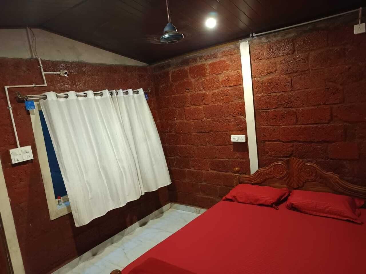 Une chambre avec un mur en briques, un cadre de lit en bois, une couvre-lit rouge, des rideaux blancs et un luminaire au plafond.