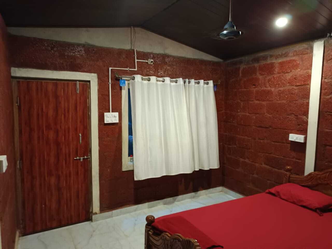 A bedroom with a red bedspread, a wooden door, a white curtain, and a brick wall.