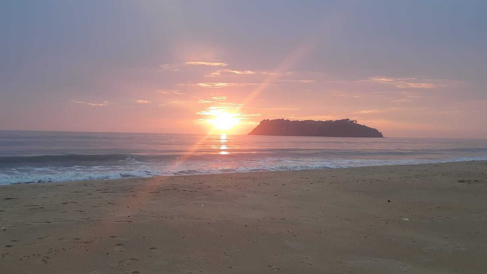 Una playa con el sol poniéndose sobre el océano, una orilla arenosa y una isla distante.