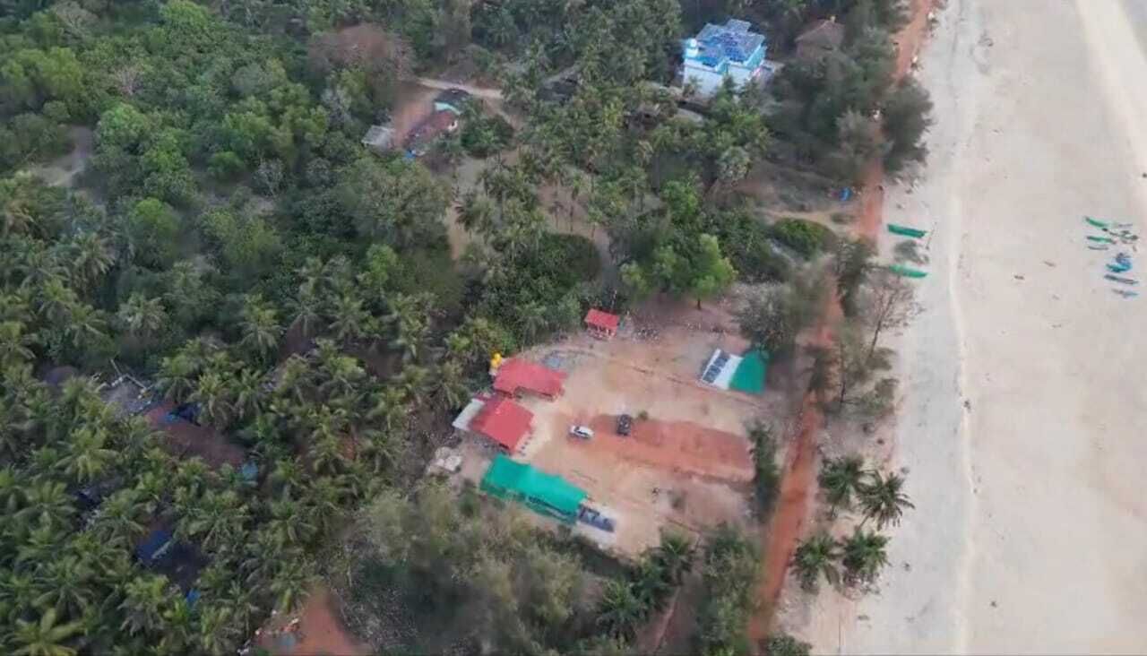 An aerial view of a coastal area with dense vegetation, a beach, and several structures with red roofs.