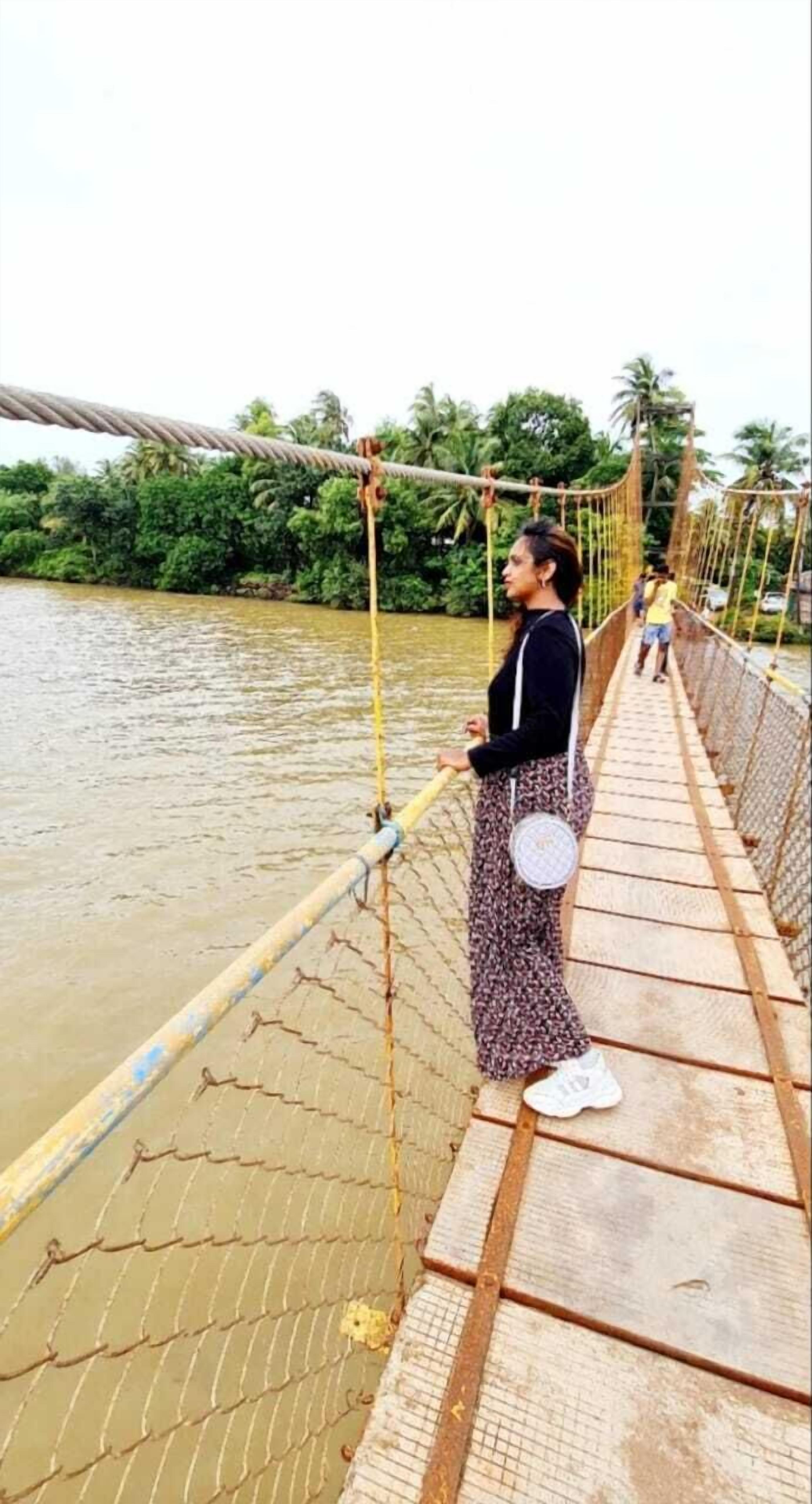 A pedestrian bridge over a river with a chain-link fence and wooden planks, surrounded by greenery.