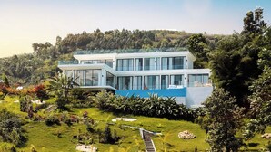Une maison moderne dotée d’une grande façade en verre, située sur une colline verdoyante et offrant une vue sur l’eau au premier plan.