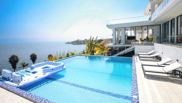 Un espace piscine moderne offrant une vue dégagée sur la mer, avec un salon aménagé sur une plateforme flottante et un bâtiment doté de grandes fenêtres.