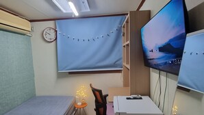 A compact room with a bed, a desk with a computer, a chair, a TV mounted on the wall, and a clock. - kinghouse (Gwangju)