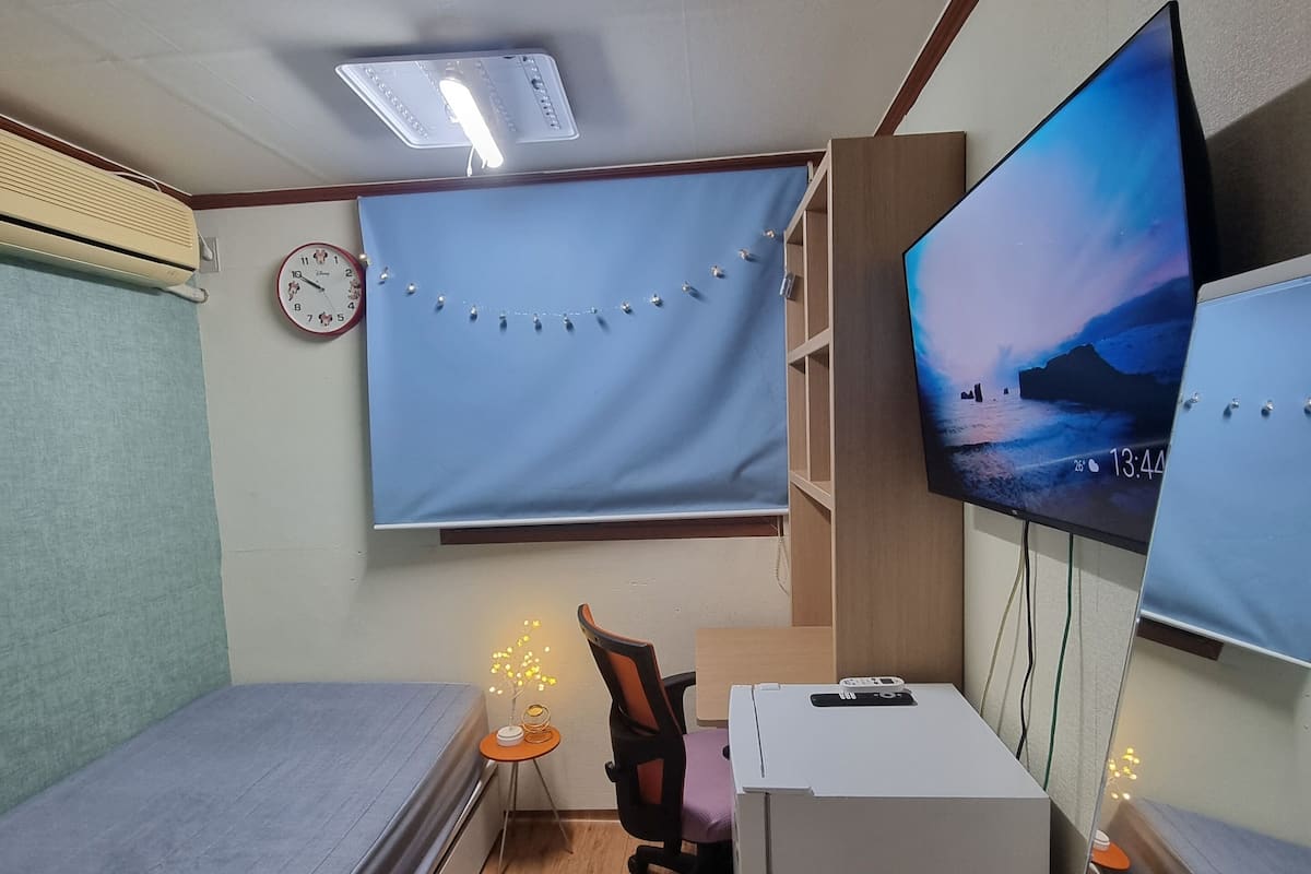 A compact room with a bed, a desk with a computer, a chair, a TV mounted on the wall, and a clock.
