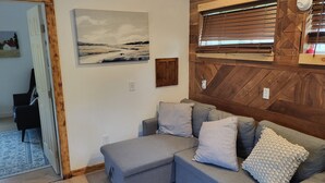 Un salon moderne avec un canapé d’angle gris, un mur décoratif en bois, un tableau et une fenêtre avec des stores.