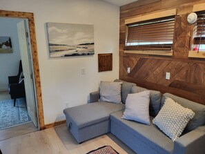 A modern living room with a grey sectional sofa, a wooden accent wall, a painting, and a window with blinds. - Terry Hill Little Lodge, Cozy, Luxury Comfort overlooking beautiful Keuka Lake! (Hammondsport)