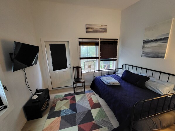 Une chambre à coucher comprenant un lit, une chaise, un téléviseur, une fenêtre avec des stores et une porte.