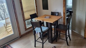Un coin repas avec une table en bois et quatre chaises, de grandes fenêtres équipées de stores et une vue sur l’extérieur.