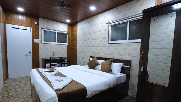 Una cama bien tendida con almohadas y una manta, un escritorio de madera con una silla, y una puerta con un ventanal.